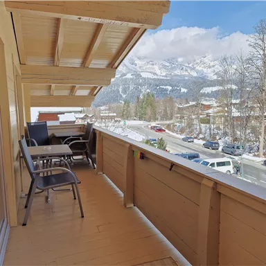 A balcony with wooden cladding, on which there are chairs and a table. In the background, snow-covered mountains and a clear blue sky can be seen.