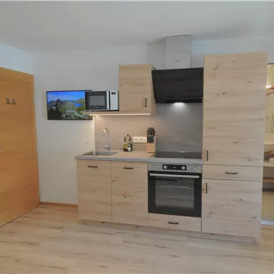 A modern kitchen unit made of wood with a stove, a refrigerator, and a microwave. In the background, a bathroom is visible, and there is a television on the wall.