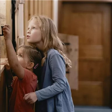 Two children are standing in a hallway and curiously looking at a wall. The girl is observing the event while the little child stands next to her.