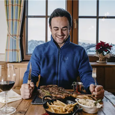 A man is sitting at a table and smiling while looking at a large piece of meat. On the table, there are also fries, salad, and a glass of red wine.
