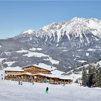 A picturesque winter location with a cozy mountain cabin. In the background, snow-covered mountains rise under a clear blue sky.