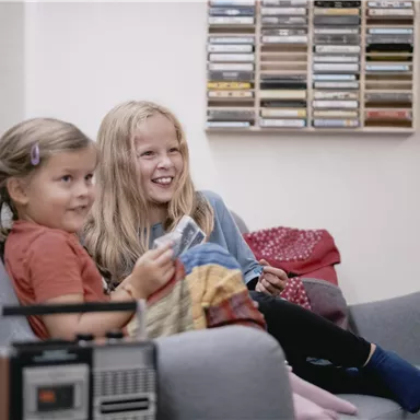 Two girls are sitting on a gray sofa and smiling. In the background, cassette tapes and a radio are visible.