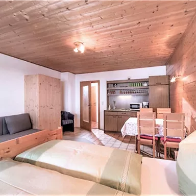 Ein gemütliches Zimmer mit Holzverkleidung, einem Doppelbett und einer kleinen Küche. Es gibt eine Sitzecke mit einer grauen Couch und einen Esstisch mit Stühlen.