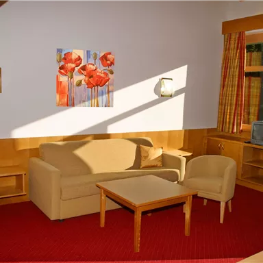 A cozy living room with a beige sofa and two chairs. A picture of red flowers hangs on the wall, and the room is well-lit.