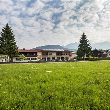A picturesque building amid a green meadow and under a cloudy sky. In the background, mountains can be seen framing the landscape.