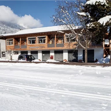 A winter landscape with a wooden building and snow-covered ground. In the background, mountains are visible under a blue sky.