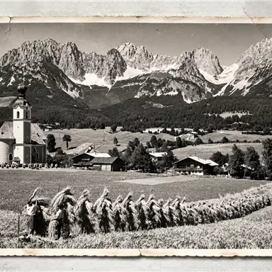 Eine nostalgische Schwarz-Weiß-Aufnahme von einem Dorf mit einer Kirche und einer beeindruckenden Berglandschaft im Hintergrund. In einem Feld ziehen Menschen in Trachten eine Reihe von Erntearbeiten.