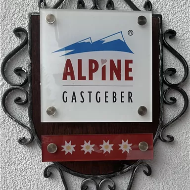 A logo with the inscription "ALPINE GASTGEBER" and stylized mountains. The colors are red, blue, and white, with Edelweiss flowers at the bottom.