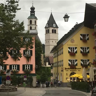 Eine malerische Stadtansicht mit bunten Gebäuden und einem hohen Kirchturm im Hintergrund. Die Straße ist von Bäumen gesäumt und es gibt Tische im Freien.