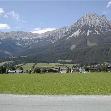 A picturesque landscape with tall mountains and green fields. In the foreground, there are some houses under a clear blue sky.