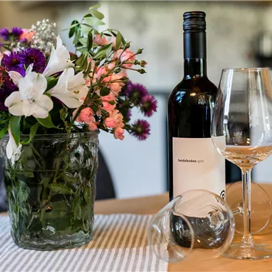 Ein Tisch mit einer Flasche Wein und zwei Weingläsern. Daneben steht ein Blumenglas mit bunten Blumen.
