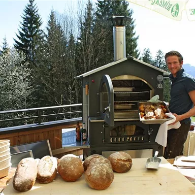 A man stands next to a wood stove, holding freshly baked bread in his hand. In the background, tall trees and a clear sky can be seen.