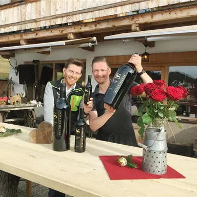 Two men are standing at a wooden table, holding wine bottles in their hands. In the background, roses and a cozy environment are visible.