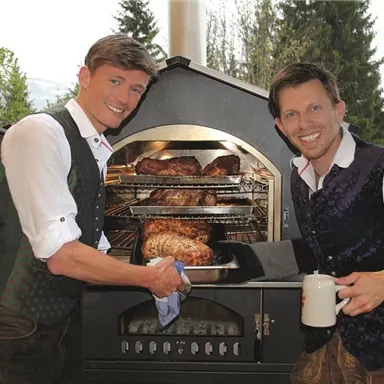 Two men in traditional clothing are standing by a grill and pointing at freshly grilled meat. The surroundings are natural, with trees in the background.