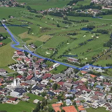Aerial view of a small village with lush green fields and a golf course. In the foreground, residential houses and a church are visible.