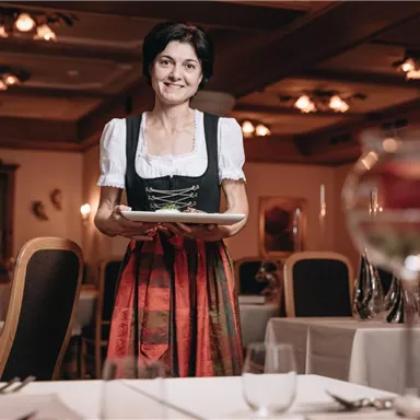 A woman in traditional attire stands in an elegant restaurant with a plate in her hand. In the background, set tables and warm light sources can be seen.