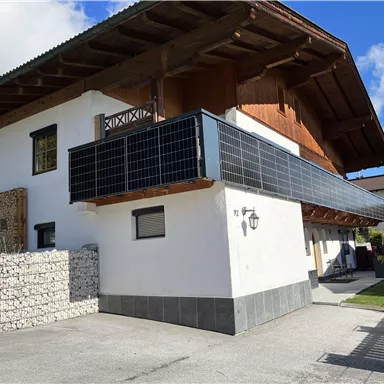 A modern house with a wooden overhang and a balcony. The facade features solar panels and there is a garden view in the background.