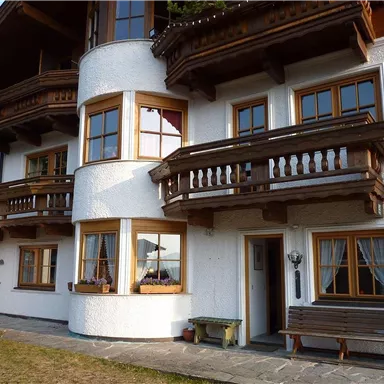 Ein charmantes, mehrstöckiges Haus im alpinen Stil mit Holzverzierungen und großen Fenstern. Es gibt Balkone und einen kleinen Gartenbereich vor dem Gebäude.