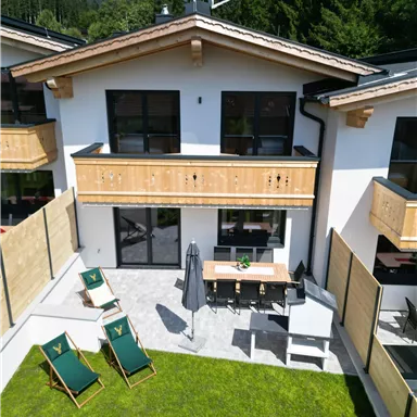 Ein modernes Chalet mit einem Garten und Terrassen. Auf der Terrasse stehen Liegestühle und ein Esstisch.