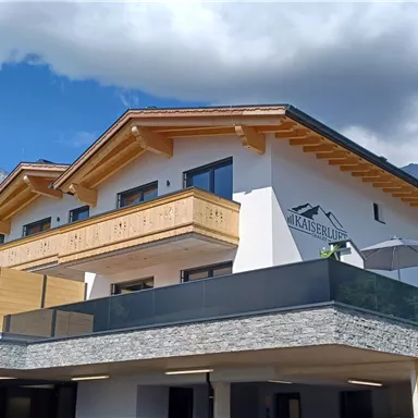 Ein modernes Gebäude in den Bergen mit Holzverkleidung und großen Fenstern. Die Umgebung ist grün und von Bäumen umgeben, unter einem klaren Himmel.