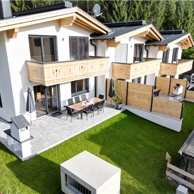 Zwei moderne Häuser mit Holzverkleidung in einer grünen Umgebung. Auf der Terrasse stehen ein Esstisch und Stühle.