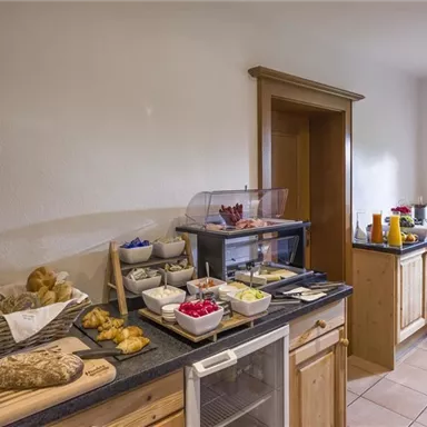A cozy kitchen with a rich breakfast selection. There are fresh rolls, fruit, and drinks on a dark table.