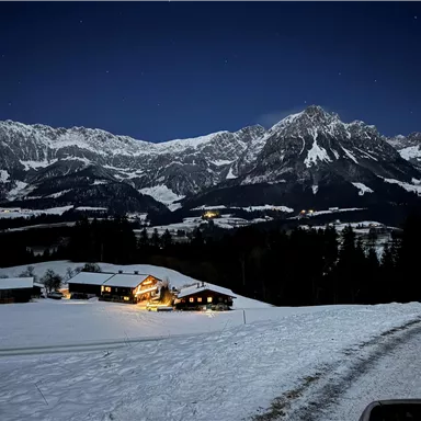 Eine verschneite Landschaft bei Nacht mit beleuchteten Häusern. Im Hintergrund erheben sich beeindruckende Berge unter einem klaren Sternenhimmel.