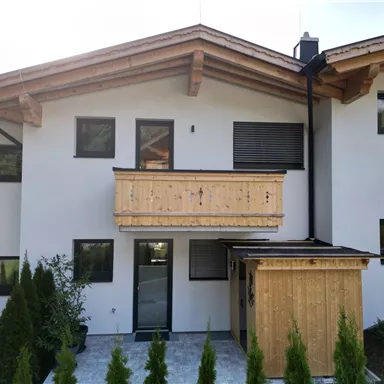 Eine moderne Fassade eines Hauses mit Holzbalustrade und großen Fenstern. Der Eingangsbereich ist klar gestaltet und von kleinen Bäumen umgeben.