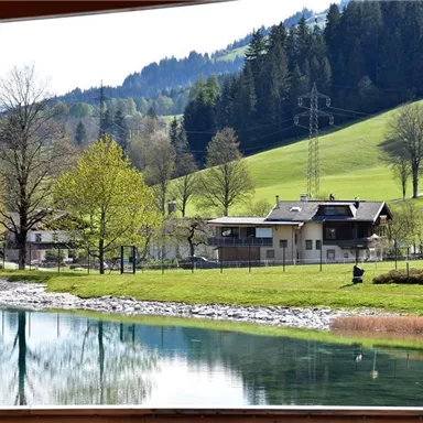 An idyllic landscape with a tranquil lake and green meadows. In the background, houses and scattered trees can be seen against a gentle hill backdrop.