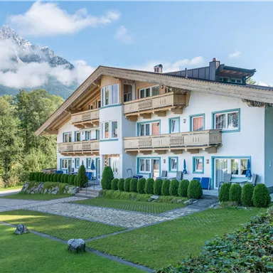 A picturesque house with wooden decorations in a green environment. In the background, mountains with clouds can be seen.