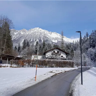 Eine verschneite Landschaft mit einem schönen Haus und hohen Bergen im Hintergrund. Der Himmel ist klar und blau.