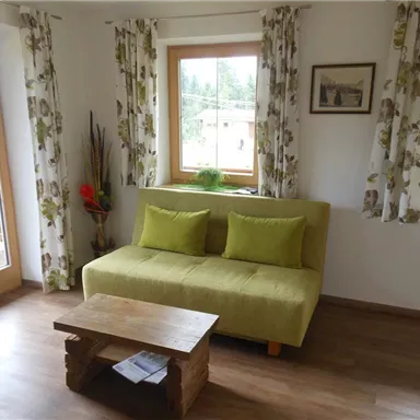 A cozy living room with a green sofa and a wooden table. Large windows let in plenty of light and there are decorative curtains.