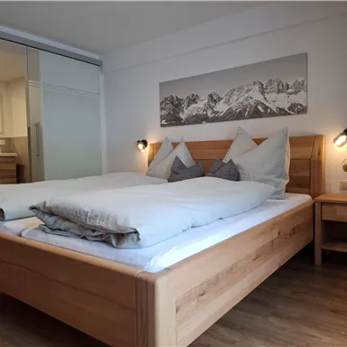 A modern bedroom with a large wooden bed and cozy pillows. A picture of mountains hangs on the wall, and there is a bright, inviting atmosphere.