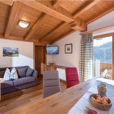 A cozy living room with a wooden beam ceiling and a dining table. Large windows provide a view of the surrounding nature.