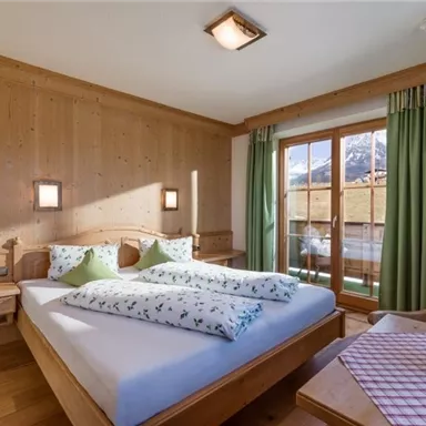 A cozy bedroom with wooden walls and large windows. The room is well-lit and has a double bed with pillows in fresh colors.