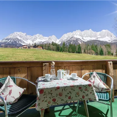 A beautiful balcony with a table and chairs, surrounded by a picturesque mountain landscape. The snow-capped mountains gleam under a clear blue sky.