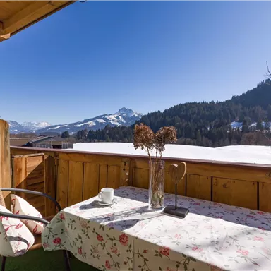 A cozy balcony with a table and chairs. In the background, a beautiful mountain landscape stretches out under a clear blue sky.