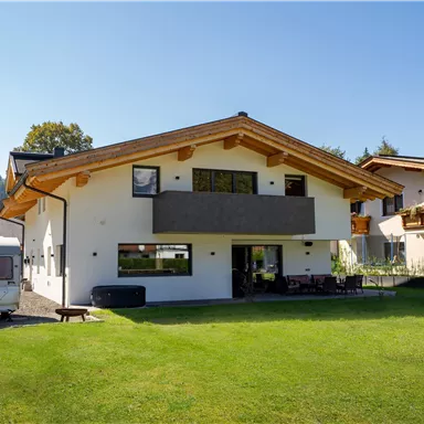 A modern house with wooden details and a large garden. In the foreground, there is a caravan and in the background are trees and meadows.