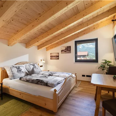 A cozy bedroom with a wooden ceiling and two beds. The room has a window overlooking the landscape and a small desk.