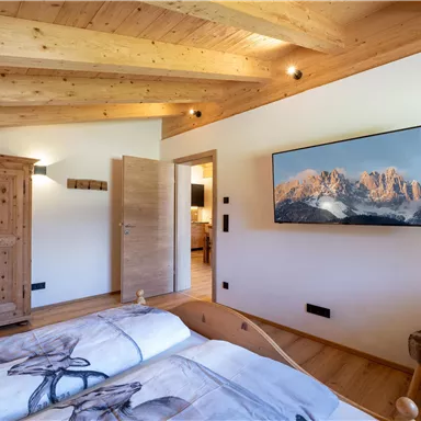 A cozy bedroom with wooden furniture and a large TV. The walls are painted light and the ceiling has visible wooden beams.