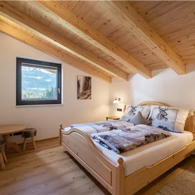 A cozy bedroom with wooden ceilings and a double bed. A window offers a view of nature and there is a small seating area.