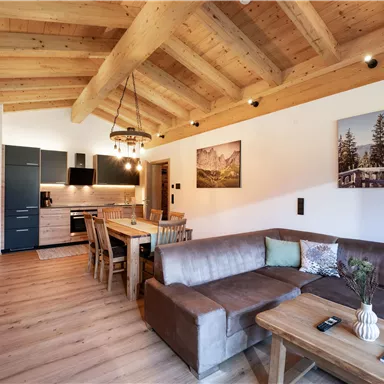 A modern living room with a wooden ceiling, a cozy couch, and a dining table. The kitchen is open and well-equipped, with large windows that let in a lot of light.