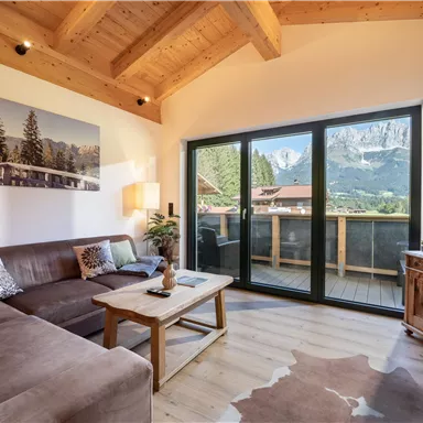 A cozy living room with a wooden ceiling and large window. From the inside, there is a view of the mountains and a terrace.