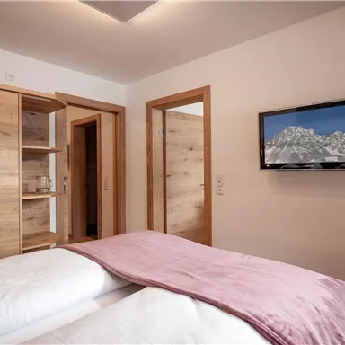 A modern bedroom with wooden cabinets and a television on the wall. The bedding is cozy and in soft colors.