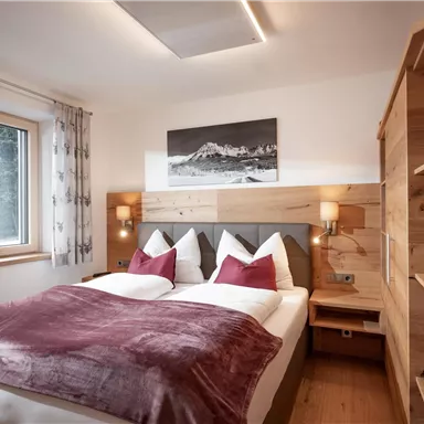 A cozy bedroom with a double bed and elegant pillows. Wooden shelves and a window create a bright atmosphere.
