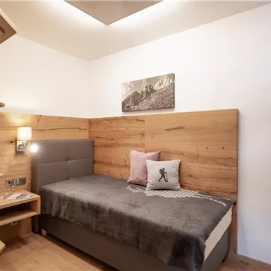 A cozy room with wooden paneling and a single bed. A television hangs on the wall, and a small window offers a view of the mountains.