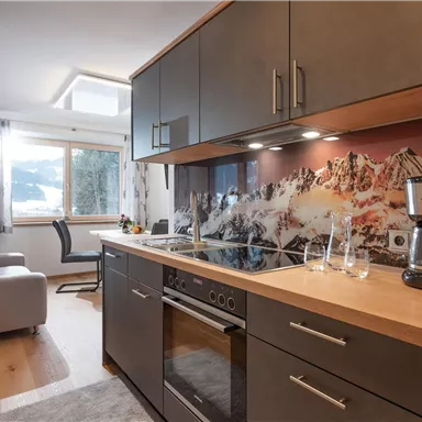 A modern kitchen with a wooden workspace and elegant cabinets. In the background, there is a cozy living area with a window and a beautiful mountain motif.