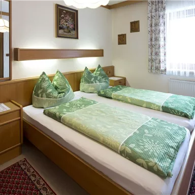 A cozy bedroom with two beds and green pillows. The room has wood paneling and large windows with light.