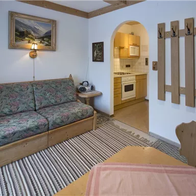 A cozy living space with a floral sofa and a dining table. The kitchen is visible through an arch and there are wooden panels on the wall.