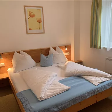 A cozy bedroom with a wooden frame bed and two large pillows. The room has a friendly ambiance with a window and a colorful picture on the wall.